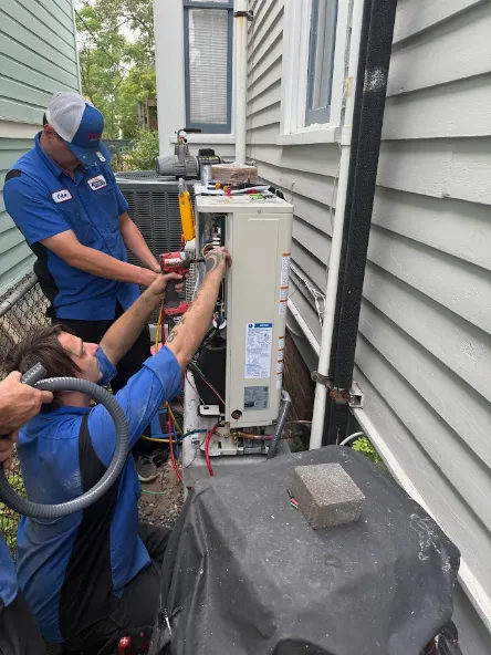 Two technicians in blue uniforms work on an outdoor HVAC unit between two houses, using tools and equipment for air conditioning repair or installation.