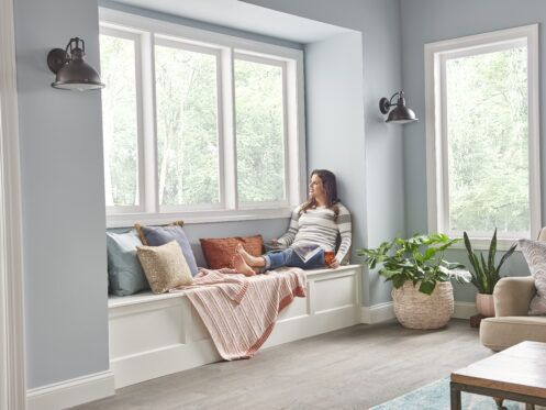 A woman sits on a window seat with pillows and a blanket, looking outside. The room has large windows, light blue walls, and a potted plant on the floor.