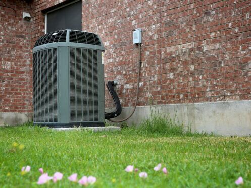 An outdoor air conditioning unit sits on a concrete slab next to a brick house, with green grass and scattered pink petals in the foreground.