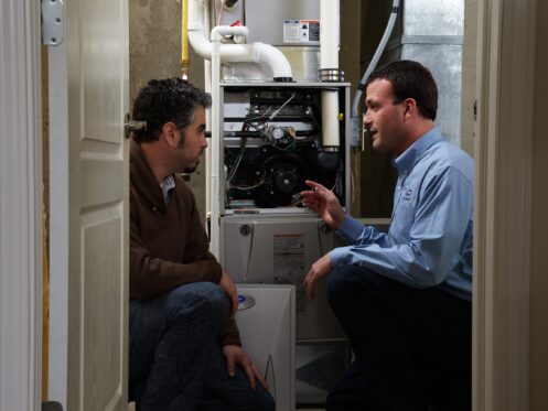 A technician in a blue shirt explains something to a man in a brown sweater while inspecting a furnace in a utility room.