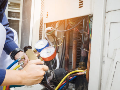 A person checks an HVAC unit using manifold gauge hoses, with wires and components visible inside the open panel.