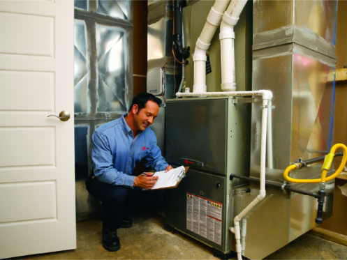 Technician kneeling by a furnace, inspecting it and writing notes on a clipboard in a utility room.
