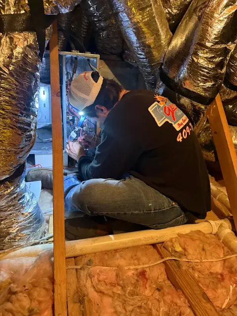 A technician in a cap and black shirt performs heating repair on an electrical unit in an attic, surrounded by insulation and ductwork.
