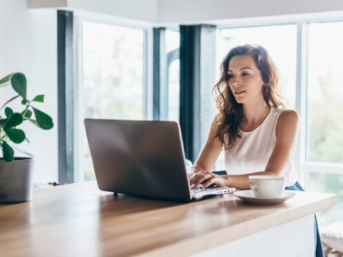 Woman sitting at a wooden table, working on a laptop with a cup and plant nearby in a bright room.