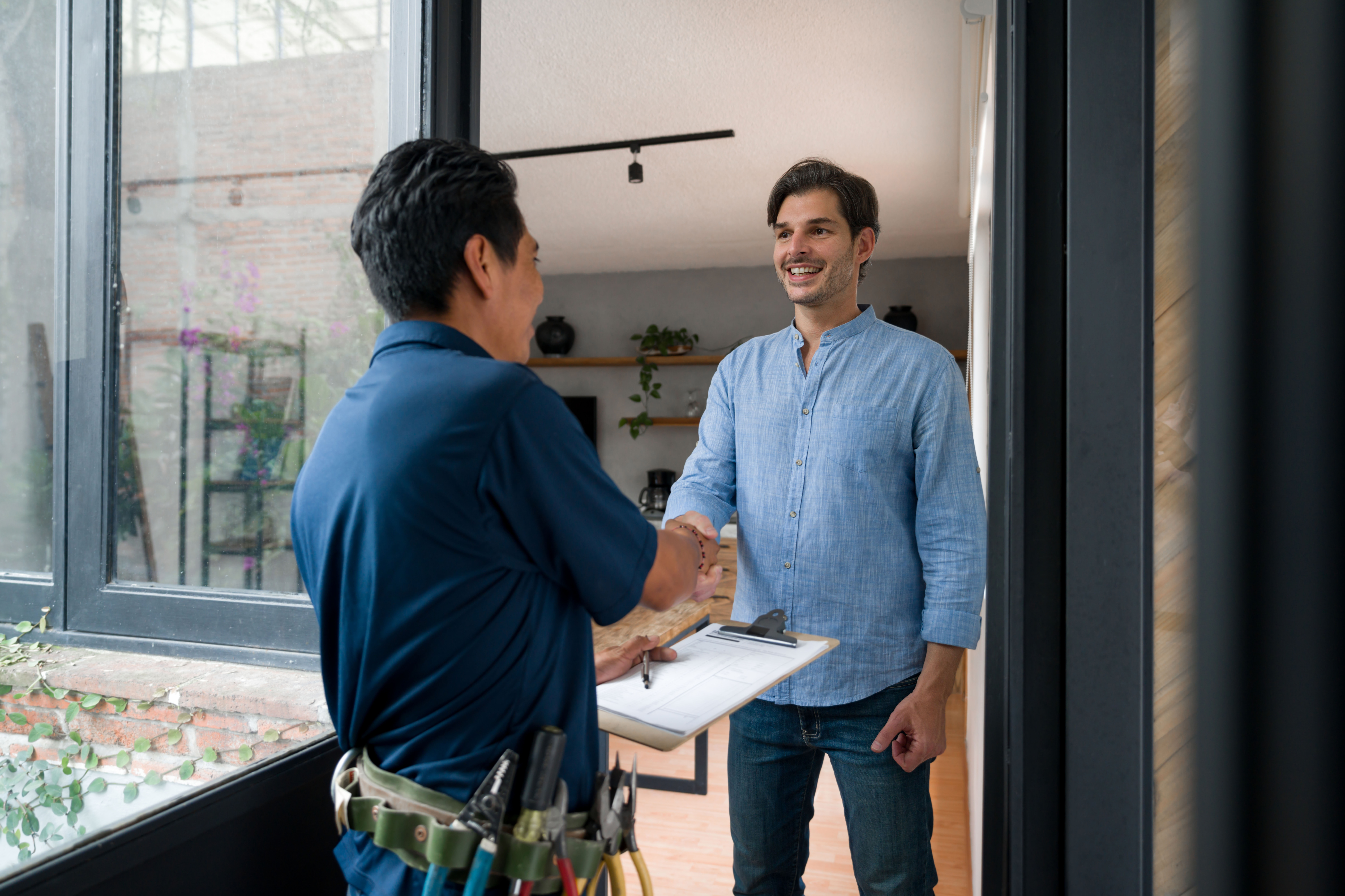 A repair technician with a clipboard shakes hands with a smiling man at the door of a modern home.